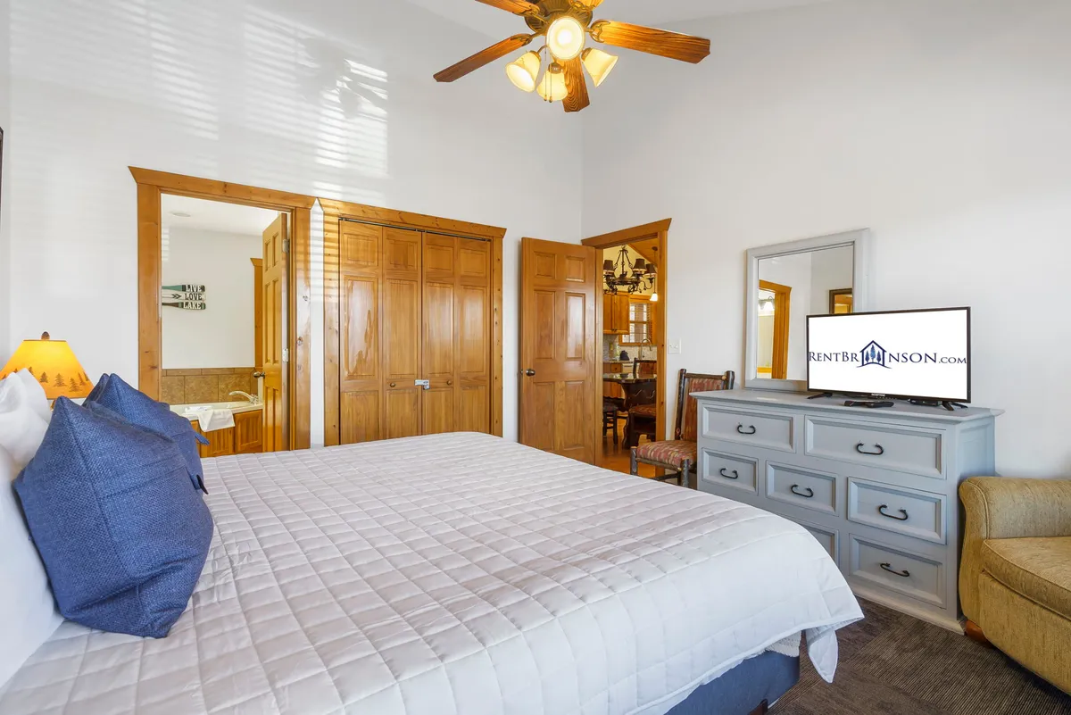 A bright, comfortable bedroom featuring a king-size bed with a white quilt and blue accent pillows, wooden furniture, and a flat-screen TV on the dresser. The room feels both cozy and luxurious, ideal for unwinding at the end of the day.