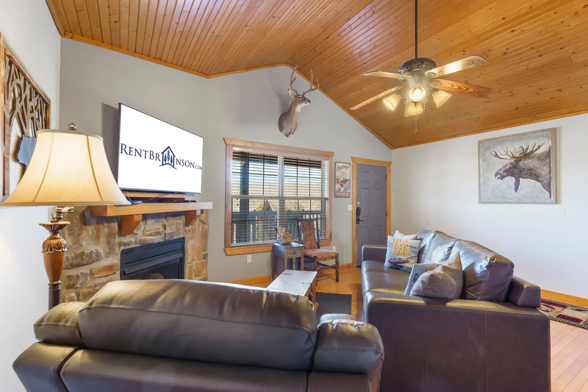 A rustic living room scene with a prominent stone fireplace, leather sofas, a wooden ceiling, a TV, and a decorative moose head on the wall. The arrangement invites relaxation, movie nights, and casual socializing in a space filled with character.