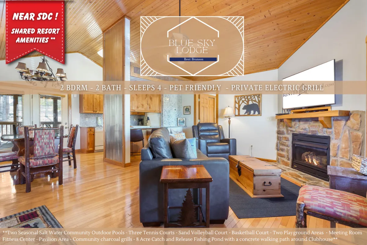 This image features an open-plan living space with a wooden ceiling, a striking stone fireplace, a comfortable seating area, and the kitchen visible in the background. The room exudes warmth and rustic charm, offering ample space for family gatherings and relaxation.