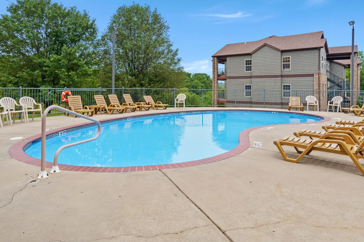 A sparkling outdoor swimming pool surrounded by lounge chairs and lush greenery, inviting guests to relax or swim under the summer sun. The pool deck is bordered by trees, creating a tranquil oasis perfect for enjoying warm weather.