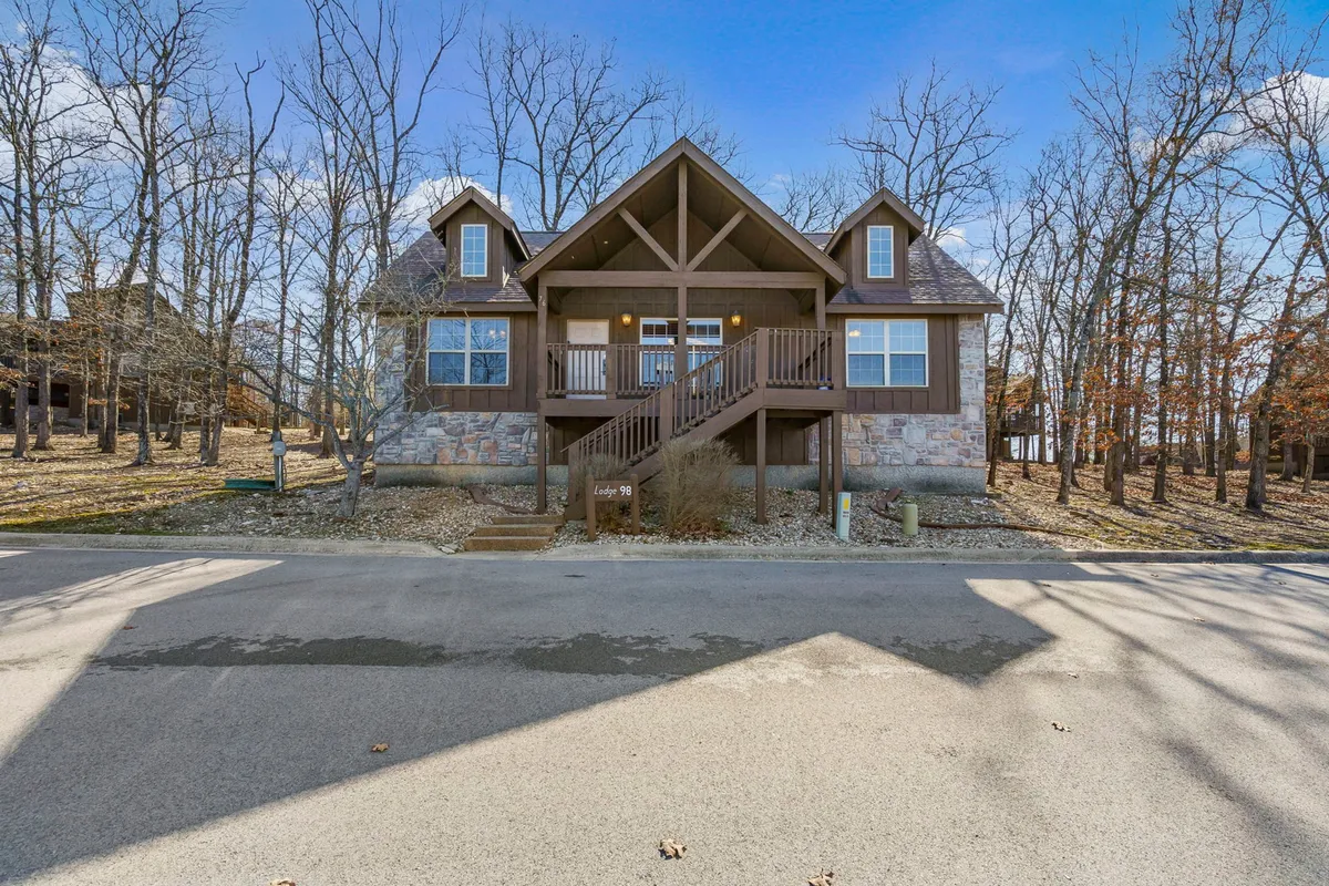 A warm exterior shot of the lodge entrance, showcasing the inviting wooden deck and stone facade. The view emphasizes cozy architecture and a sense of welcome, with the surrounding trees enhancing the natural, serene environment.
