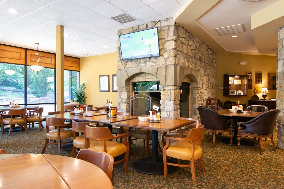 An inviting interior view of the Ledgestone Grille, highlighting a stone fireplace, cozy tables and chairs, a television, and large windows. The atmosphere is relaxed and social, ideal for unwinding after a round of golf.