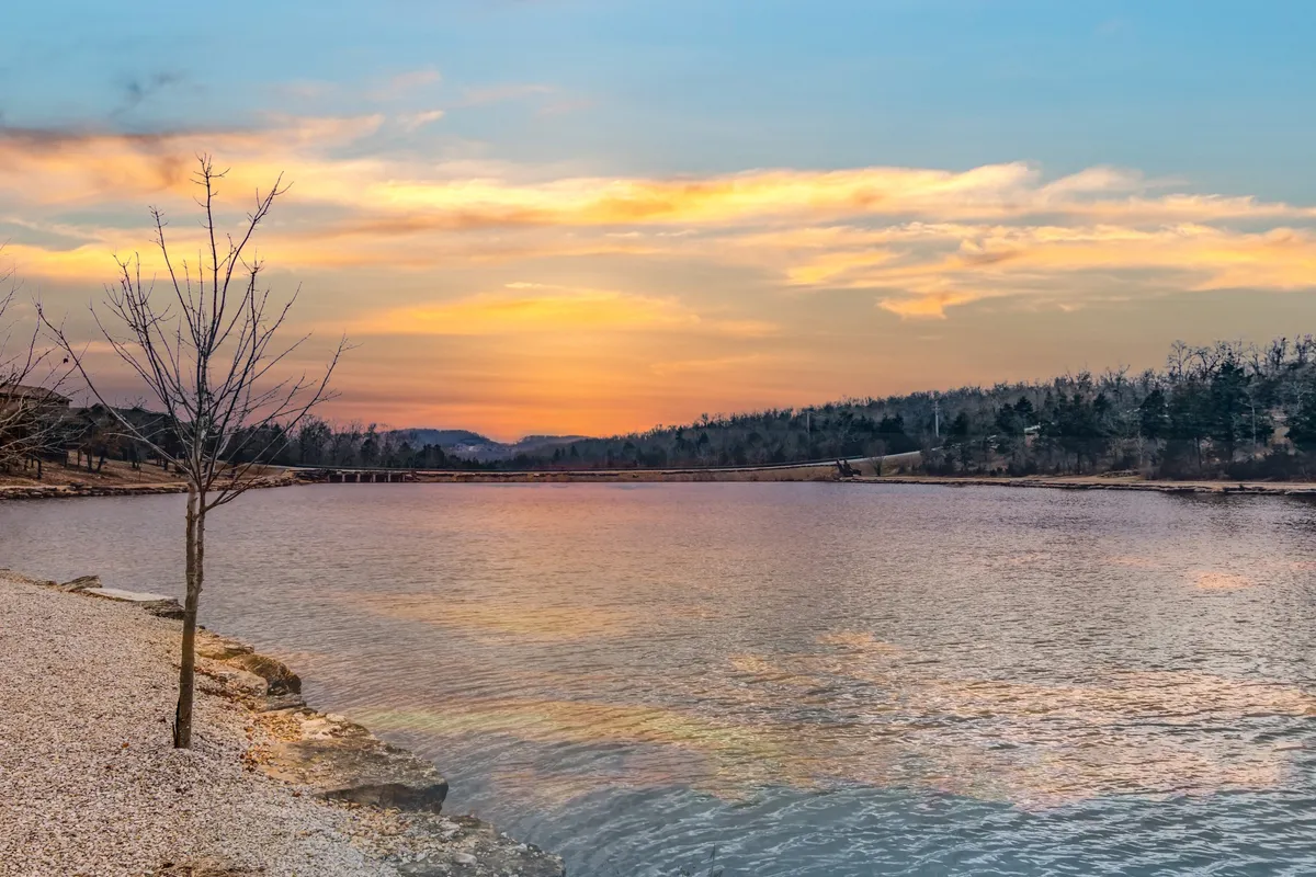 A tranquil lake scene at sunset, with the water reflecting warm colors and a tree framing the view. The distant hills provide a scenic backdrop, while the lakeshore is inviting for fishing, walking, or picnicking.