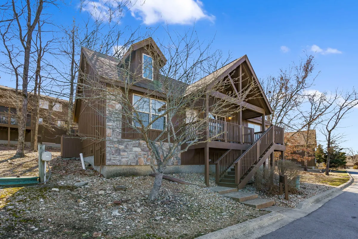 This exterior image showcases a wooden deck attached to a stone-facade lodge, surrounded by mature trees. The deck is furnished for gatherings and embraces the natural setting, offering a seamless transition from indoor comfort to the outdoors.