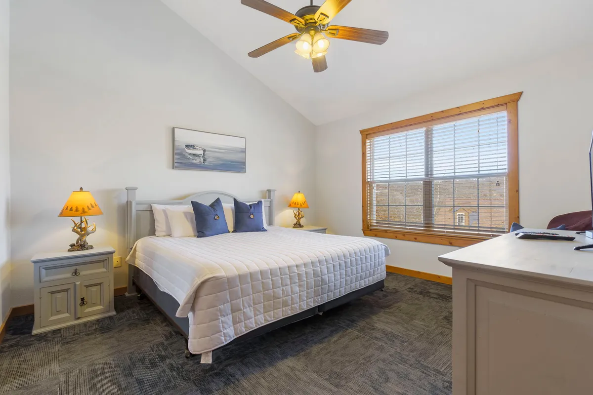 A tranquil bedroom featuring a large bed with plush linens, soft lighting from nightstands, a window allowing in natural light, and a ceiling fan above. The decor is neutral and calming, with a focus on comfort and relaxation.
