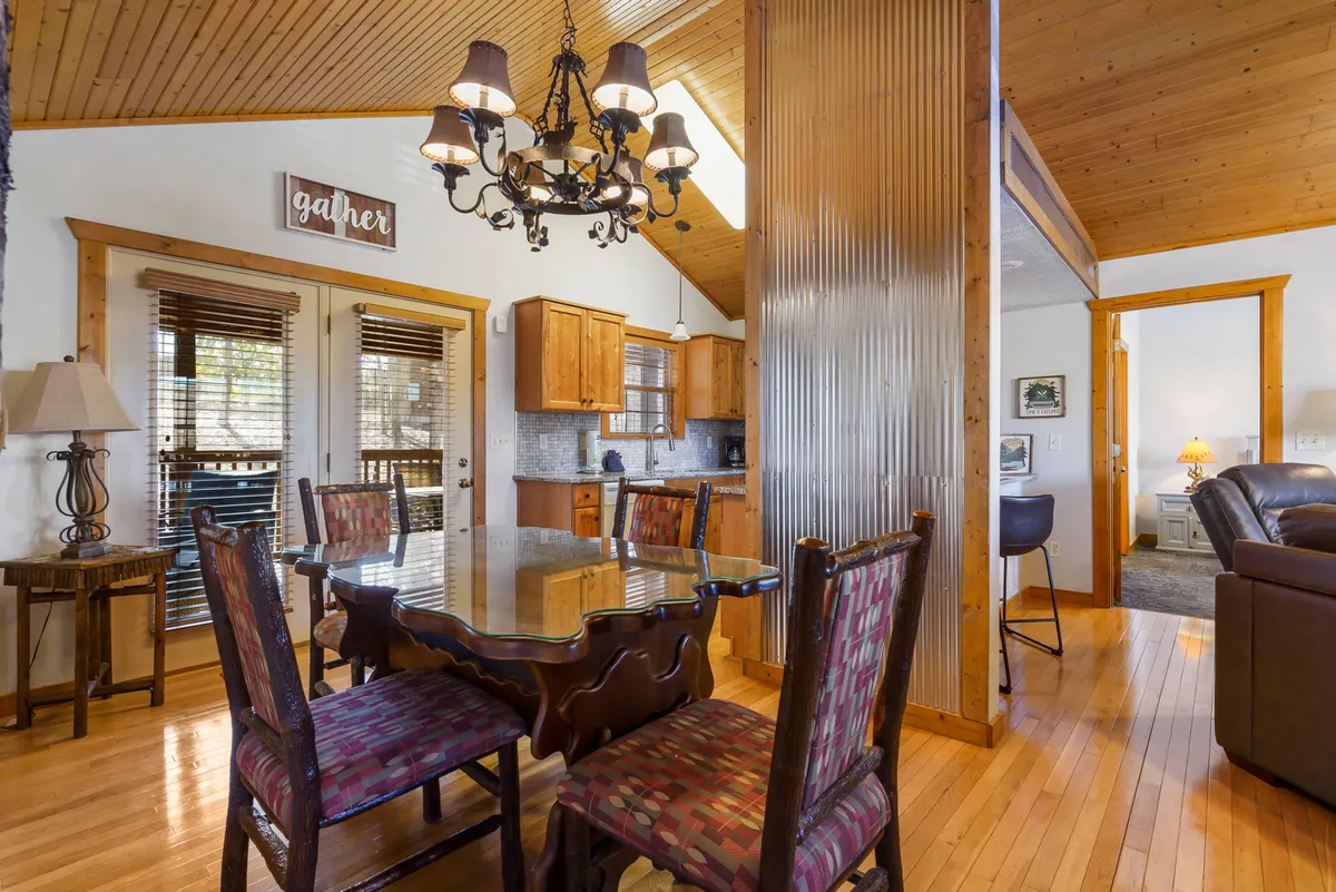The image highlights a welcoming dining table beneath a chandelier, with the kitchen area and exposed wooden beams visible in the background. A decorative 'gather' sign hangs above, reinforcing the communal spirit of the space.