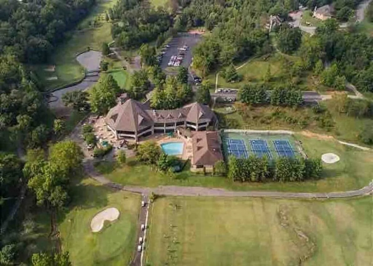 A wide exterior shot that encompasses the main resort building, swimming pool, tennis courts, and a glimpse of the golf course, all nestled within a lush landscape. The image conveys the scale of the resort’s amenities and their proximity to the lodging and golf course.