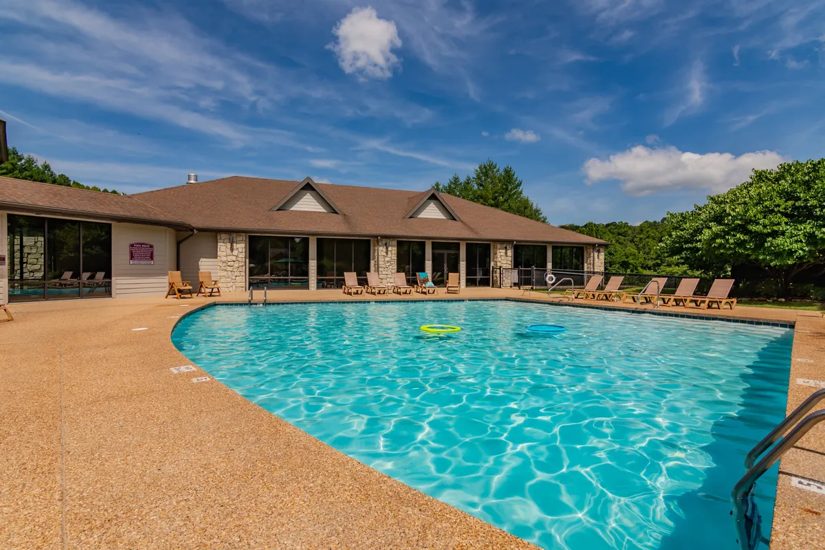 A sparkling blue pool surrounded by lounge chairs and lush greenery, with a building exterior in the background. The area is inviting for swimming, sunbathing, or relaxing in the shade during warm months.
