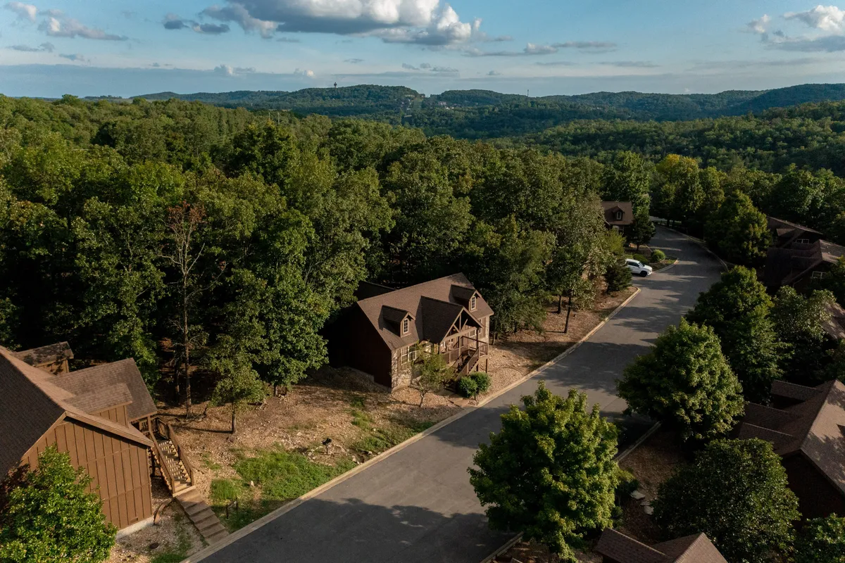 A relaxing exterior view of multiple wooden cabins enveloped by vibrant greenery, paved walking paths, and distant mountain views. The scene hints at outdoor gatherings, hiking, and peaceful mornings spent on the porch.