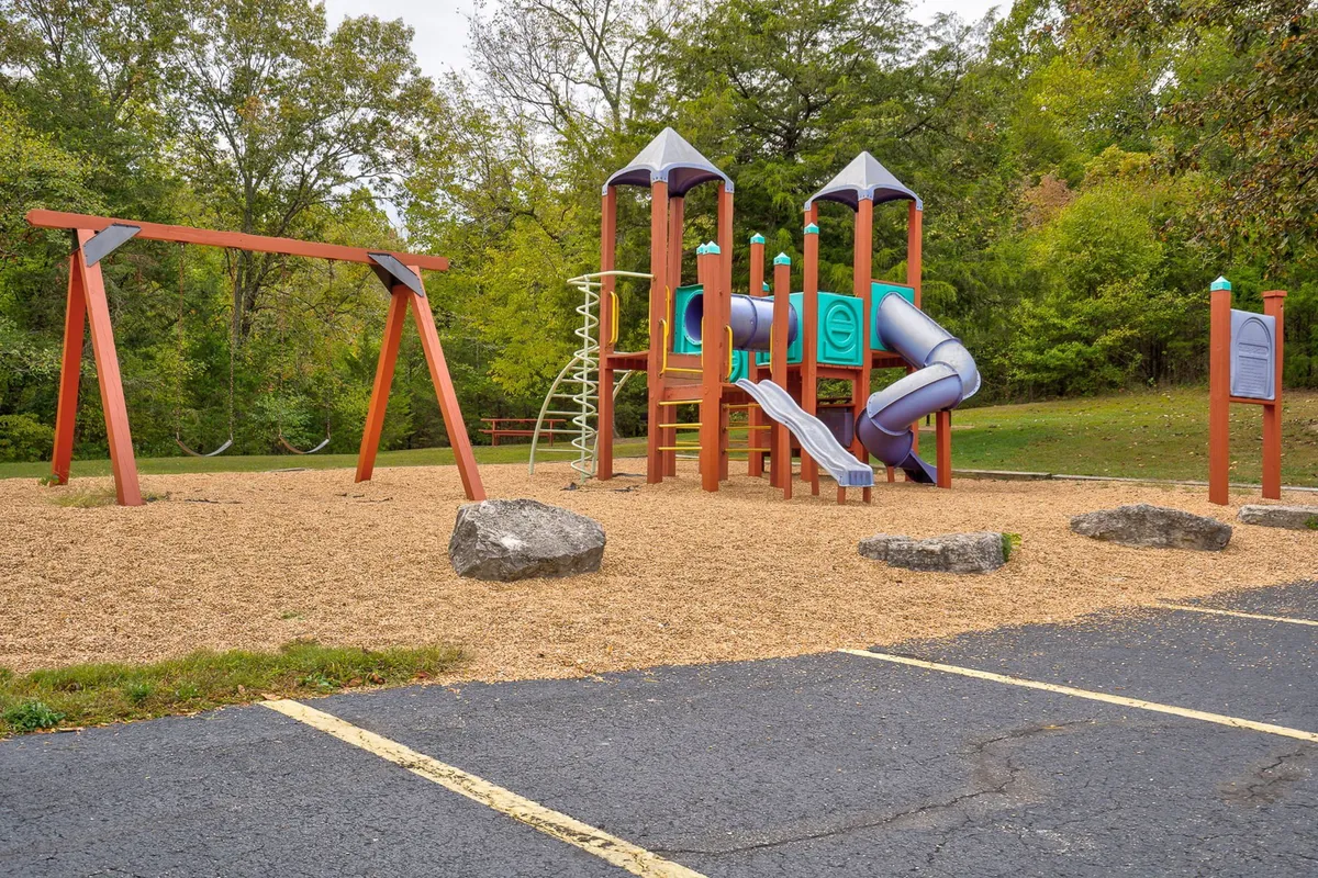 A vibrant playground set among trees, featuring swings, rocks for climbing, and open space for children to play. The area is surrounded by greenery, creating a safe and inviting environment for kids and families.