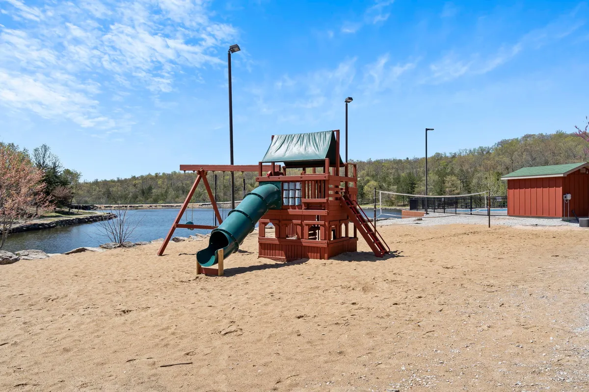 A vibrant playground scene with slides and a sand area set beside a calm lake and surrounded by green trees. Families and children enjoy outdoor fun, while the scenic water view adds a sense of relaxation and connection to nature.